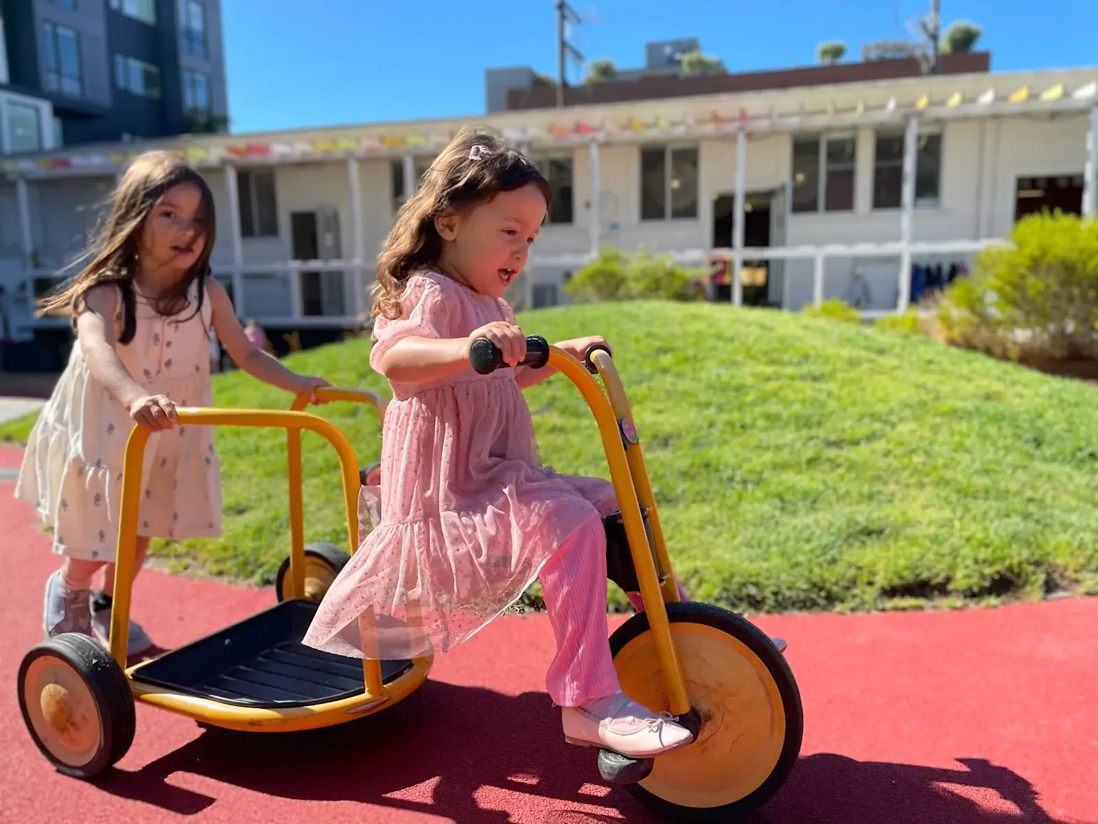 Children riding a yellow trike past the grassy mound in the Dogpatch play yard