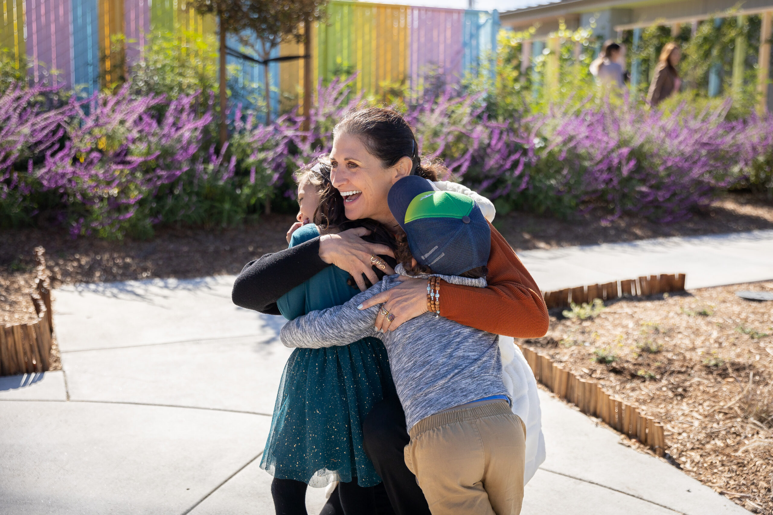 A teacher hugging two children on a garden path lined with purple blooms