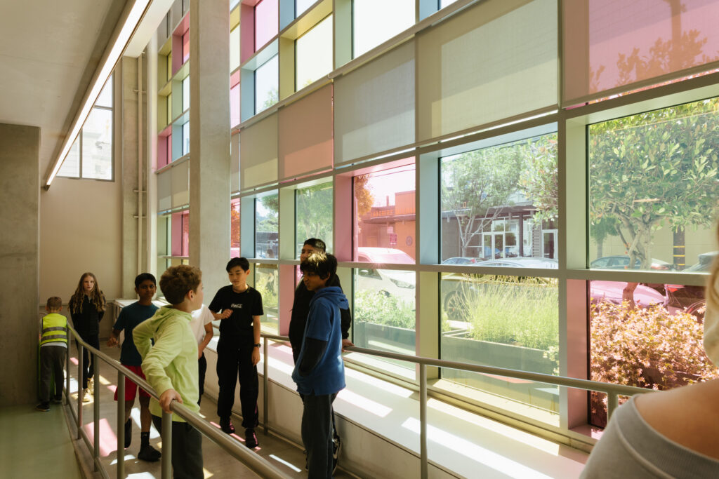 Students gathered along a sunlit walkway lined with panes of colored glass