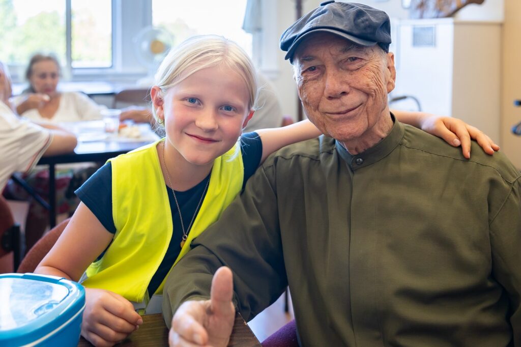 A La Scuola student in a yellow vest gives a thumbs-up beside a smiling grandfather during a community visit