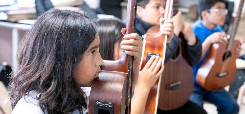 A student playing the ukulele in the music atelier with classmates behind her