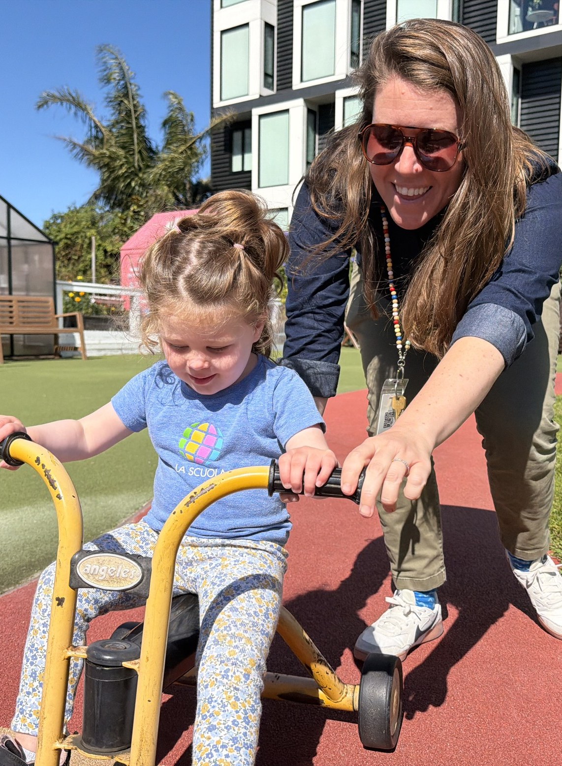 Mallory Matthews steadies a preschooler riding a yellow tricycle on the Dogpatch campus playground.