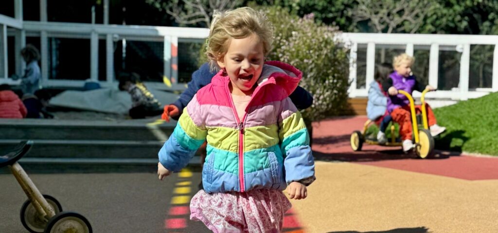 Preschooler in a rainbow jacket running across the painted Dogpatch yard during camp