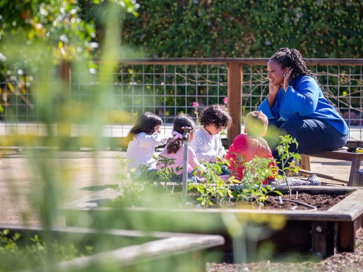 A La Scuola teacher crouches beside preschoolers planting in the raised garden beds at the Dogpatch campus.