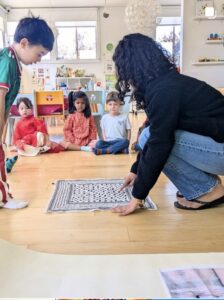 A teacher laying out a floor game while children wait, seated in a circle