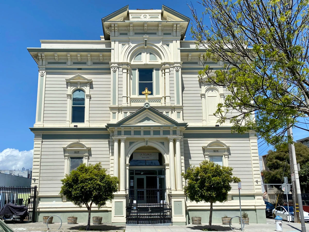 Historic white Italianate building with an arched entrance and young trees along the pavement