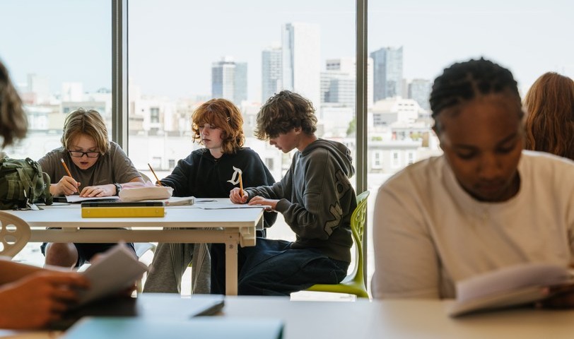 Students reading and writing at a long table in front of the skyline windows