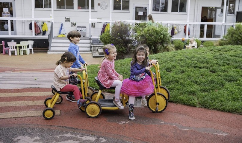 Children riding trikes past the grassy mound in the preschool yard