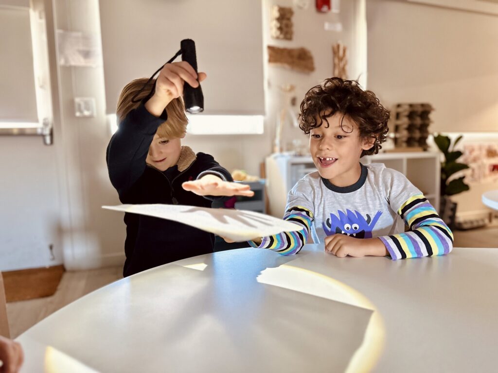Two children experimenting with a flashlight and paper at an atelier table