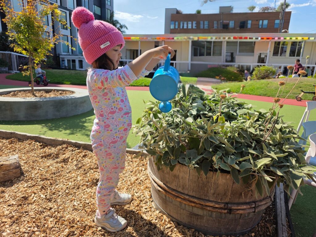 A preschooler in a pink hat watering plants in the garden