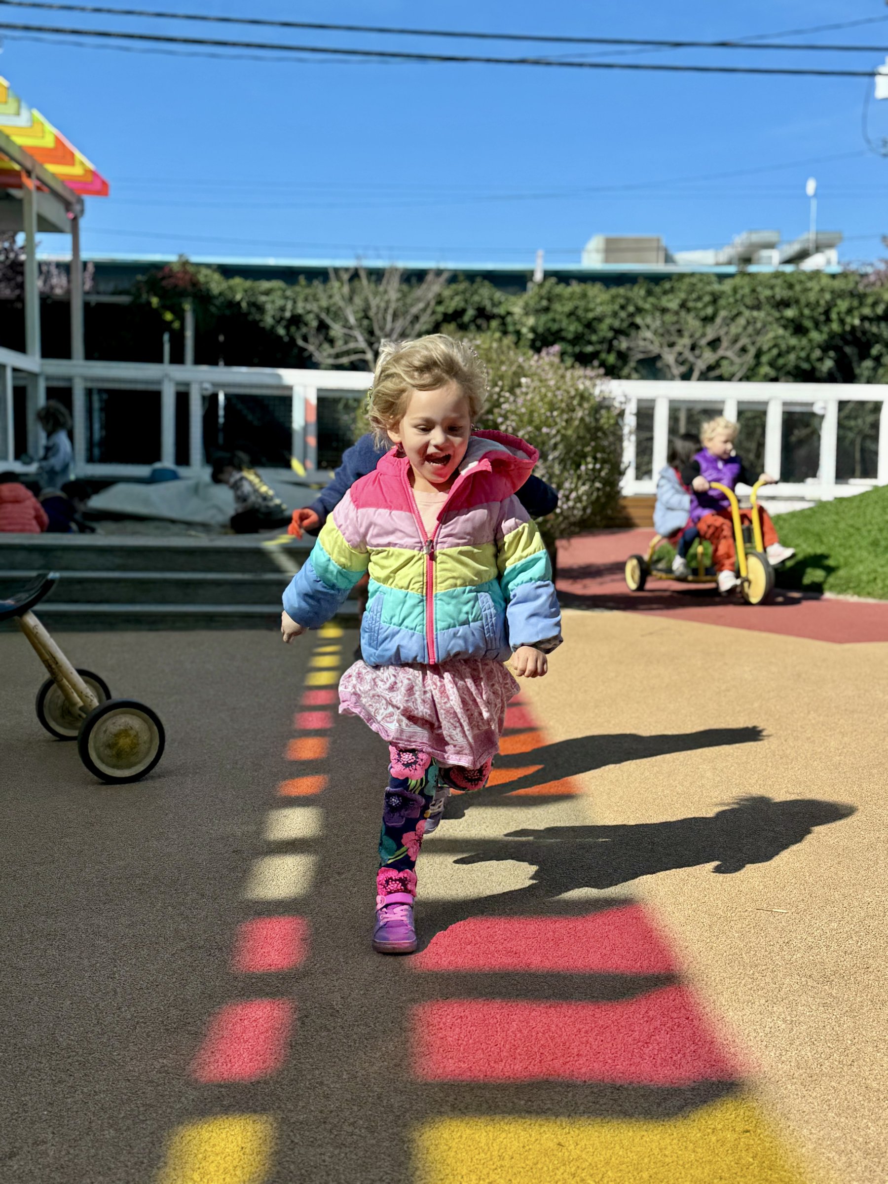 A preschooler in a rainbow jacket running across the Dogpatch campus play yard