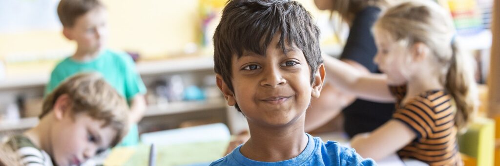 A smiling elementary student in a Superman shirt writing at his desk