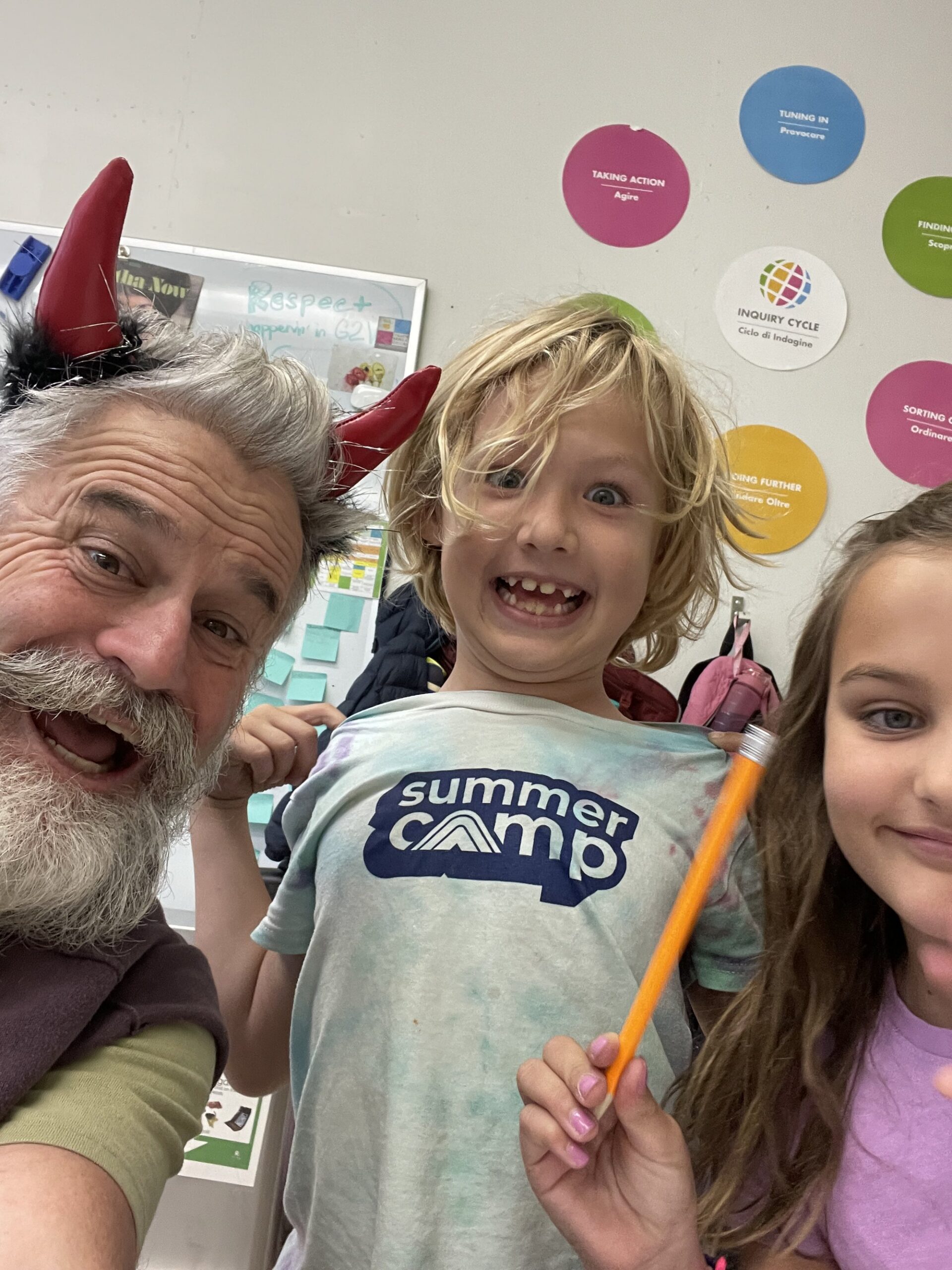 A camp counselor in a costume headband taking a selfie with two smiling campers, one in a summer camp T-shirt