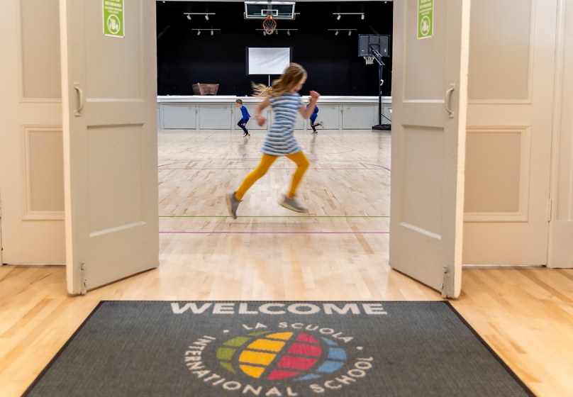 A student running across the gym floor seen through the open doors and the La Scuola welcome mat